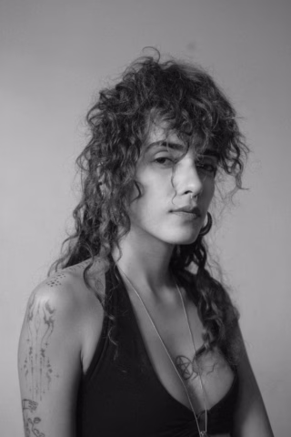 Black and white portrait of a person with curly hair, wearing a sleeveless top, hoop earrings, and a necklace. Visible tattoos are on their arm and chest. The person looks at the camera with a neutral expression.