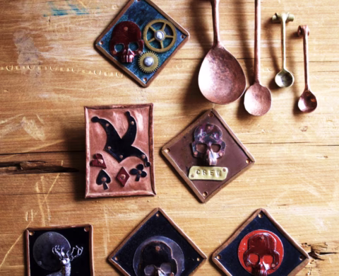 Handcrafted metal art pieces and four vintage measuring spoons are arranged on a wooden surface. The art features skulls, gears, and playing card symbols in various colours and shapes.