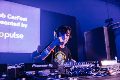 A young boy wearing headphones and a black T-shirt stands at a DJ booth, smiling and making a shaka hand sign. Bright stage lights and a large screen with event text are visible in the background.