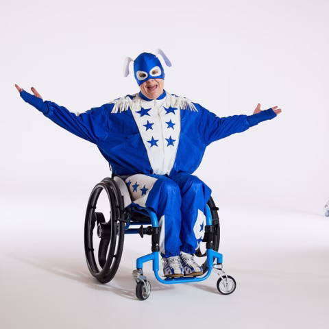 A person in a blue and white star-patterned superhero costume sits in a wheelchair, smiling with arms outstretched against a plain white background.