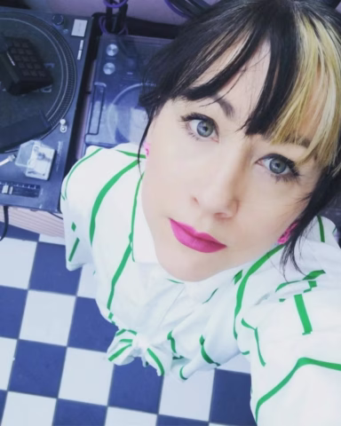 A woman with light skin, bright pink lipstick, and black hair with a blonde streak looks up at the camera. She wears a white shirt with green stripes, standing on a black-and-white chequered floor near DJ equipment.