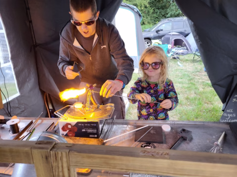 An adult and a child wearing safety spectacles work with a flame and glass rods at a table inside a marquee, possibly participating in a glassblowing demonstration at an outdoor event.