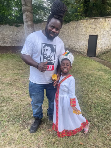 A man wearing a white graphic T-shirt and jeans stands on grass next to a smiling young girl dressed in a white traditional outfit with a headwrap. They are outdoors near a stone wall and trees.
