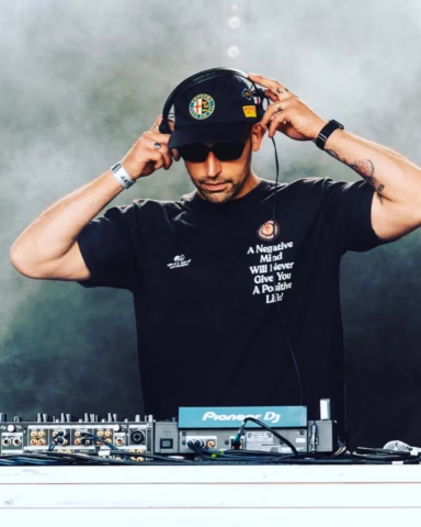 A DJ wearing sunglasses, a black cap, and a black T-shirt stands behind a Pioneer DJ mixer, adjusting his headphones with both hands. Smoke or fog creates a dramatic background.