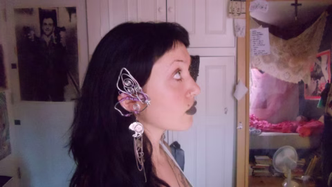 A woman with long dark hair and black lipstick is in profile, wearing an elaborate wire ear cuff. The background shows a cluttered bedroom with posters, handwritten notes, and unmade bed visible.