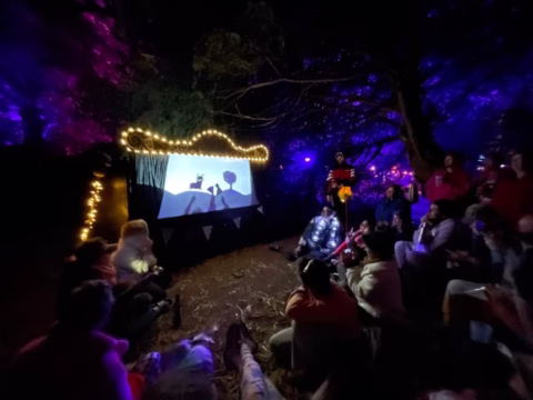 A group of people sit on the ground at night, watching a shadow puppet show illuminated by fairy lights in a forest setting with purple and blue ambient lighting.