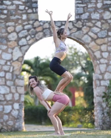 Two women perform an acro yoga pose outdoors; one woman stands bent over while the other balances on her back with arms raised. They are framed by a stone arch and surrounded by greenery.