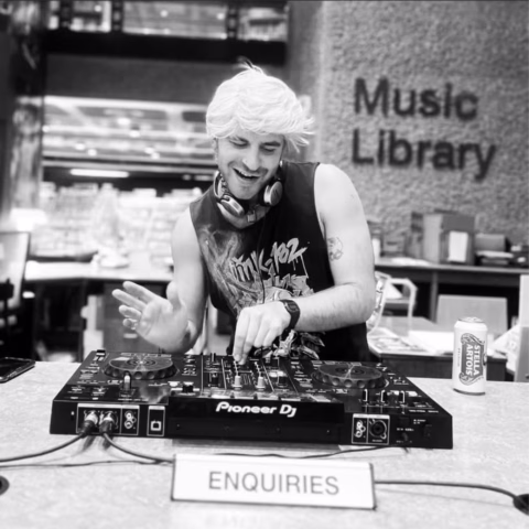 A smiling person with fair hair DJs at a Pioneer DJ deck on a desk labelled “ENQUIRIES” in a library, with headphones around their neck. A drinks can and shelves of books are visible in the background. The sign reads Music Library.