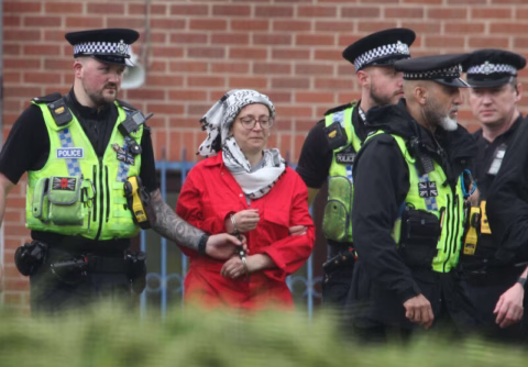A woman in a red coat and headscarf is being escorted by four police officers in yellow vests and hats, with a brick building in the background.