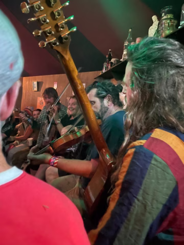 A group of musicians play string instruments closely together in a lively indoor setting, surrounded by people. The scene is colourful and energetic, with bottles and decorations visible in the background.
