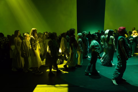 A large group of people stands and walks on a dark stage, illuminated by dramatic yellow and green lighting, creating deep shadows and highlights across the scene.