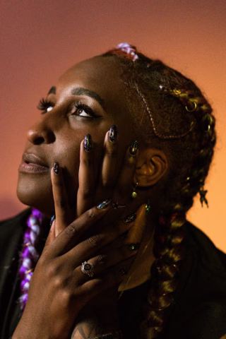 A person with plaited hair, decorated with beads, gazes upward. Their hands, featuring long, decorated nails and rings, gently touch their face. The background is softly lit in warm tones.
