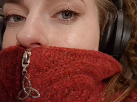 Close-up of a person wearing black headphones and a red zip-up jumper with a Celtic knot zip pull, covering their mouth. Only the upper part of their face, including one eye, is fully visible.
