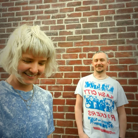 Two people stand in front of a brick wall. The person on the left, with short blond hair, is smiling and looking down, wearing a blue shirt. The person on the right has short hair and a beard, wearing a white graphic T-shirt.