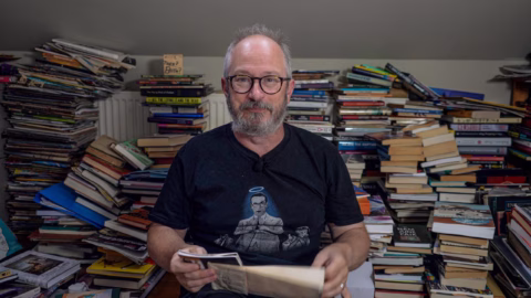 A man with glasses and a grey beard sits holding papers, surrounded by tall stacks of books in a cluttered room. He wears a black T-shirt featuring a graphic of a person with a halo over their head.