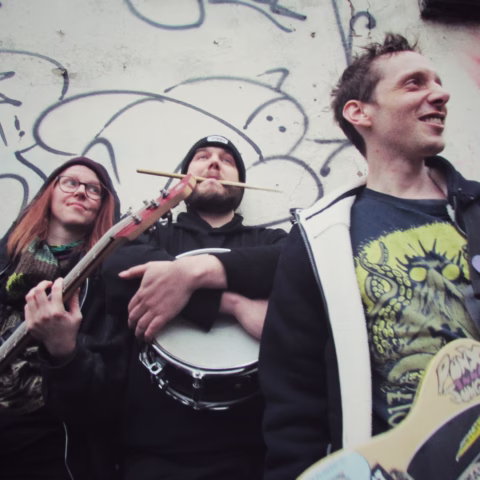Three people with musical instruments stand in front of a graffiti-covered wall. One holds a guitar, another a snare drum with drumsticks in his mouth, and the third smiles, wearing a graphic T-shirt.