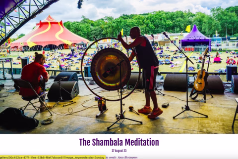 Two musicians are on an outdoor stage at a festival. One is playing a large gong while the other sits with a guitar. Colourful tents and people relaxing on the grass are visible in the background.