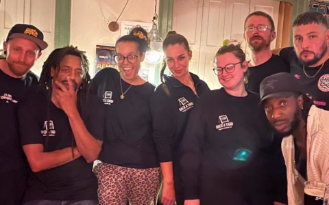 A group of seven people pose together indoors, most wearing black shirts with BACKYARD logos. They are smiling and standing closely, some with arms round each other, in a warmly lit room.