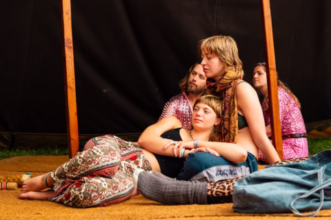 Two people sit on the ground under a wooden structure, one leaning against the other with eyes closed. Two more people sit behind them in patterned clothing. A blue bag and socks are in the foreground.