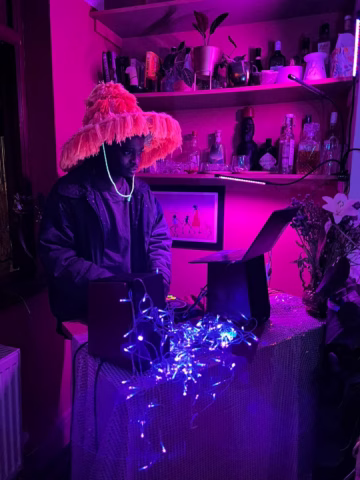 A person wearing a large, fluffy hat and glow necklace stands at a table with a laptop and speakers, surrounded by purple lights and bottles on shelves in a dimly lit, neon-pink room.