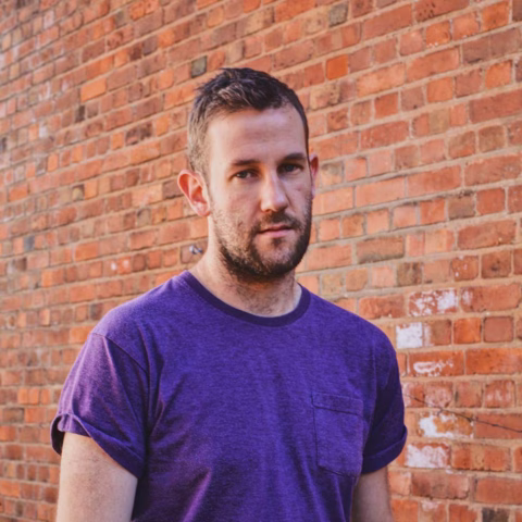 A man with short brown hair and a beard, wearing a purple T-shirt, stands in front of a red brick wall, looking at the camera with a neutral expression.