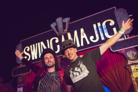 Two men in hats stand smiling with arms raised in front of a brightly lit sign that reads SWINGAMAJIG. The background features red curtains and decorative elements, suggesting a festive or performance event.