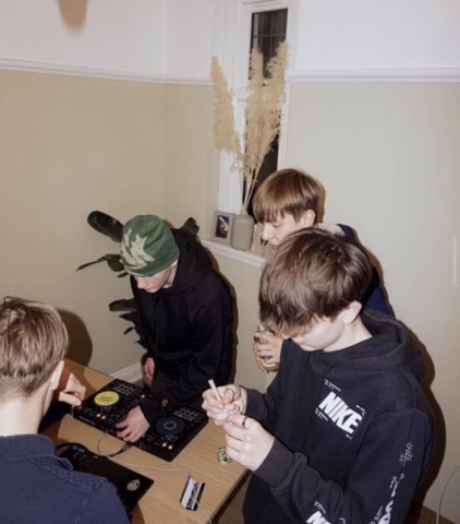 Four young people stand around a table with DJ equipment in a room with beige walls and a potted plant. One person holds a lighter and paper, another wears a green hat, and two focus on the turntables.
