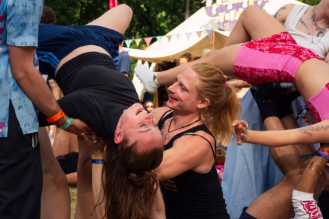 A woman laughs joyfully whilst being held upside down by others at an outdoor festival, with colourful tents and bunting in the background. Several people are participating in playful acrobatic activities.