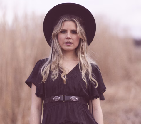 A woman with long blonde hair wearing a wide-brimmed black hat and a black dress stands outdoors in front of tall, blurred grass.