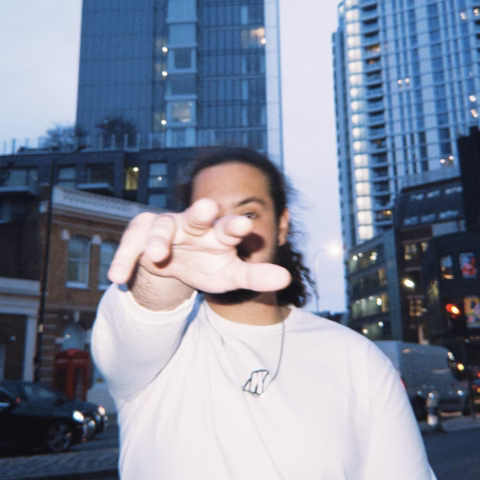 A person with long hair wearing a white shirt stands outdoors in a city, reaching towards the camera with an outstretched hand, partially obscuring their face. Tall buildings rise in the background during dusk.