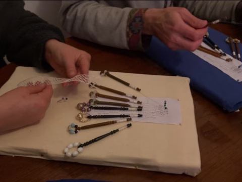 Two people work on lace-making with bobbins and pins on a pillow, their hands arranging threads to create an intricate lace pattern on a wooden table.
