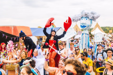 A lively outdoor festival crowd, with people in colourful costumes. One person wears a police hat and large red lobster claws, and another holds a giant cloud puppet. Tents and festive decorations are visible in the background.