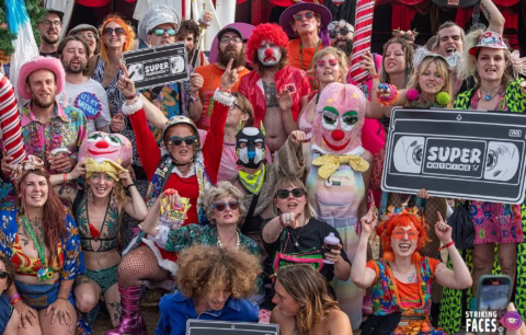 A large group of people in colourful, eclectic costumes and wigs pose together outdoors, many smiling and holding “Super” signs. The scene is lively and festive, with playful and clown themes throughout.