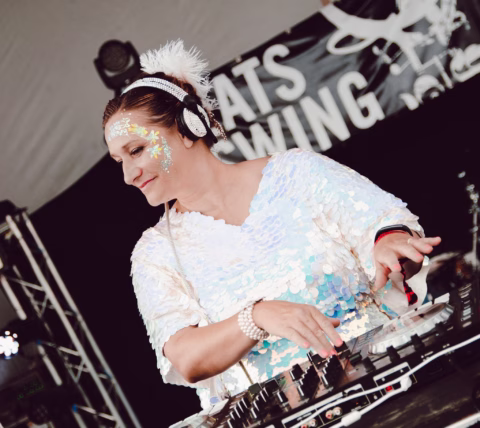 A woman wearing headphones and a sequinned outfit with face glitter is DJing at an event, adjusting controls on a DJ mixer. A KATS SWING sign is visible in the background.