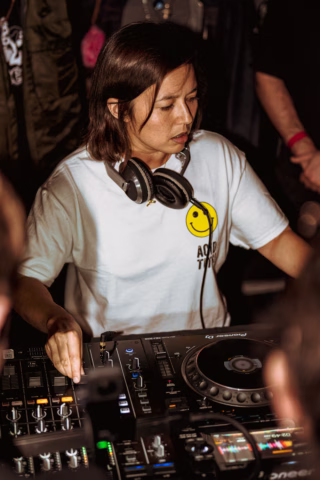 A DJ wearing a white T-shirt with a yellow smiley face and headphones round her neck mixes music on a DJ controller at a crowded event.