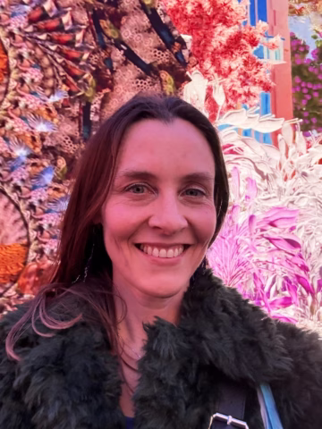 A woman smiling and taking a selfie in front of a bright, colourful, and intricate floral background with vivid red, pink, and green patterns. She is wearing a dark, textured jacket and has long brown hair.