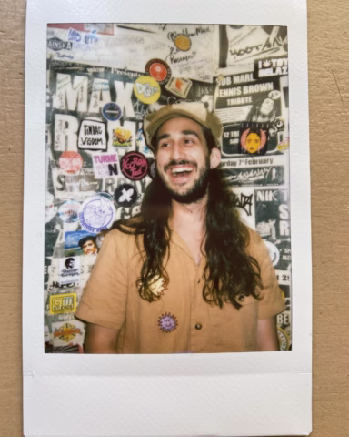 A person with long dark hair and a beard, wearing a tan shirt and matching cap, smiles whilst standing in front of a wall covered with colourful posters and stickers. The photo is framed as an instant film print.