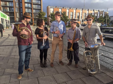 Five people stand outdoors on a paved walkway near water and modern buildings, holding brass instruments and wearing casual clothes with hats. They appear to be chatting and smiling in the sunlight.