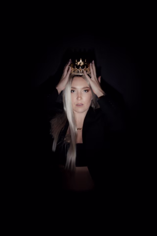 A woman with long blonde hair, wearing a black outfit, holds a gold crown on her head. She is centred against a dark background, with dramatic lighting highlighting her face and crown.