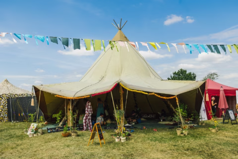 A large beige teepee tent stands on grass under colourful pennant flags at a festival, with people inside and potted plants at the entrance. Other tents and a blue sky are visible in the background.