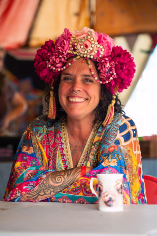 A smiling person with curly dark hair wears a colourful floral headdress and a bright patterned robe. Their arms are tattooed, and they sit at a table with a mug featuring a dog’s face. The background is festive and colourful.