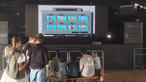 A group of children and adults sit and stand in front of a large screen displaying a retro video game character selection menu inside a dark marquee-like venue.