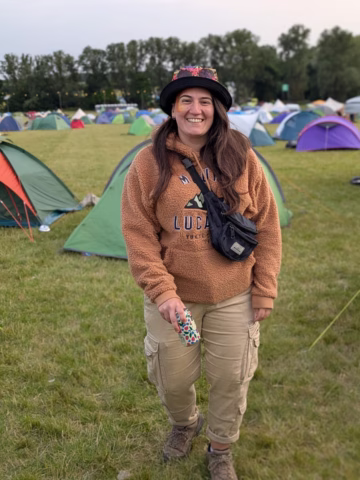 A person smiles whilst standing on grass at a campsite, surrounded by colourful tents. They wear a brown fleece, cargo trousers, a black crossbody bag, and a hat with flowers, and hold a can with a colourful design. Trees are in the background.