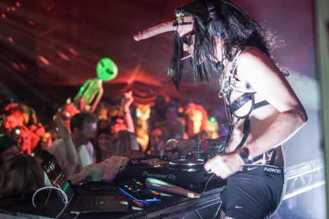A DJ wearing a fancy dress with a long nose, sunglasses, headphones, and chains performs at a lively party. The crowd behind includes someone holding an inflatable green alien. The scene is colourful and energetic.