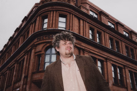 A man with curly hair and a beard, wearing a brown blazer and light shirt, stands in front of a historic red-brick building with arched windows on a cloudy day.