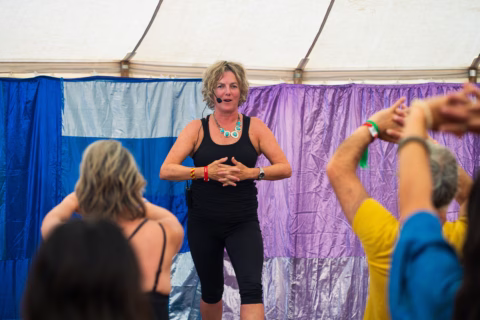 A woman in black workout clothes leads a fitness or dance class onstage in front of a colourful curtain, while participants follow her movements with arms raised.