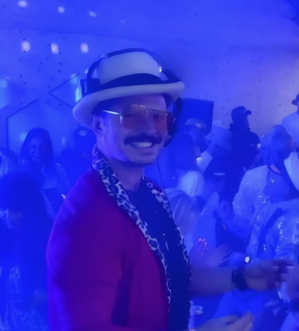 A man in a hat, sunglasses, and red jacket smiles and poses at a lively party with blue lighting, surrounded by people dancing and socialising in the background.