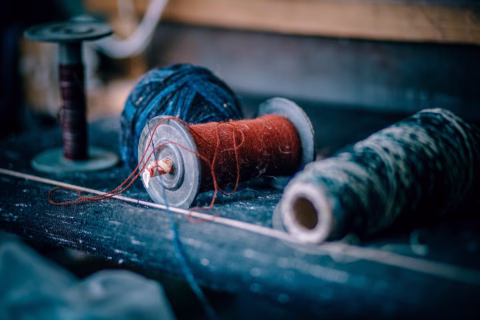 Close-up of three reels of thread in different colours—red, blue, and a mix of black and white—lying on a textured dark surface, with threads loosely unravelling from the reels.