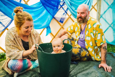 A smiling baby sits in a green tub, with a woman and a man sitting nearby inside a tent decorated with blue fabric. The adults look at the baby with joy.