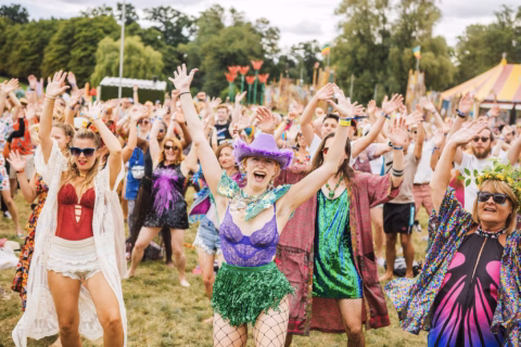 A large group of people at an outdoor festival raise their hands and smile, dressed in colourful and festive outfits, with a circus-style tent and trees visible in the background.